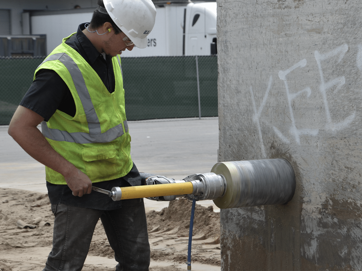 A worker in a yellow safety vest expertly operates a core drilling machine on a concrete wall at the bustling construction site.